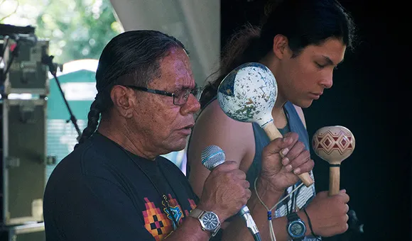Two men with long hair, one older and one younger, perform with eyes closed on stage. They each hold a painted rattle, and the older man in the foreground also holds a microphone.