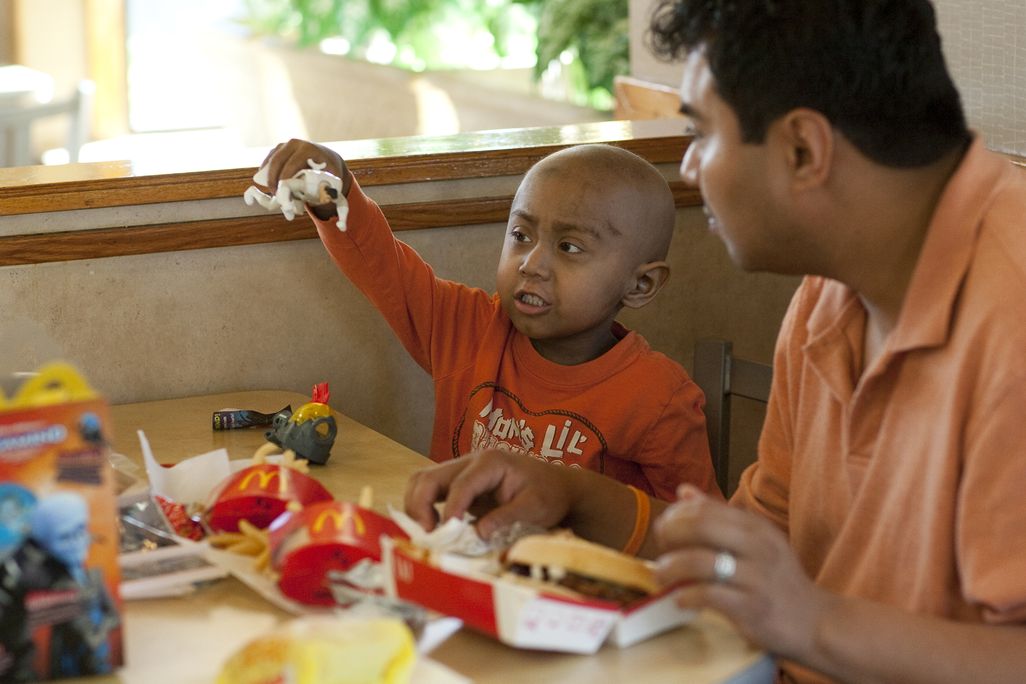 An image of a 5-year old child playing with a McDonald's Happy Meal toy. In front of the child is a happy meal with fries and seated next to the child is his father.