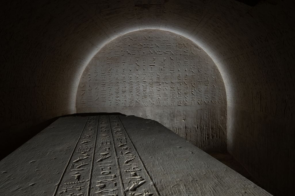 the top of a sarcophagus covered in writing with a glyph-covered wall behind it lit in an arch