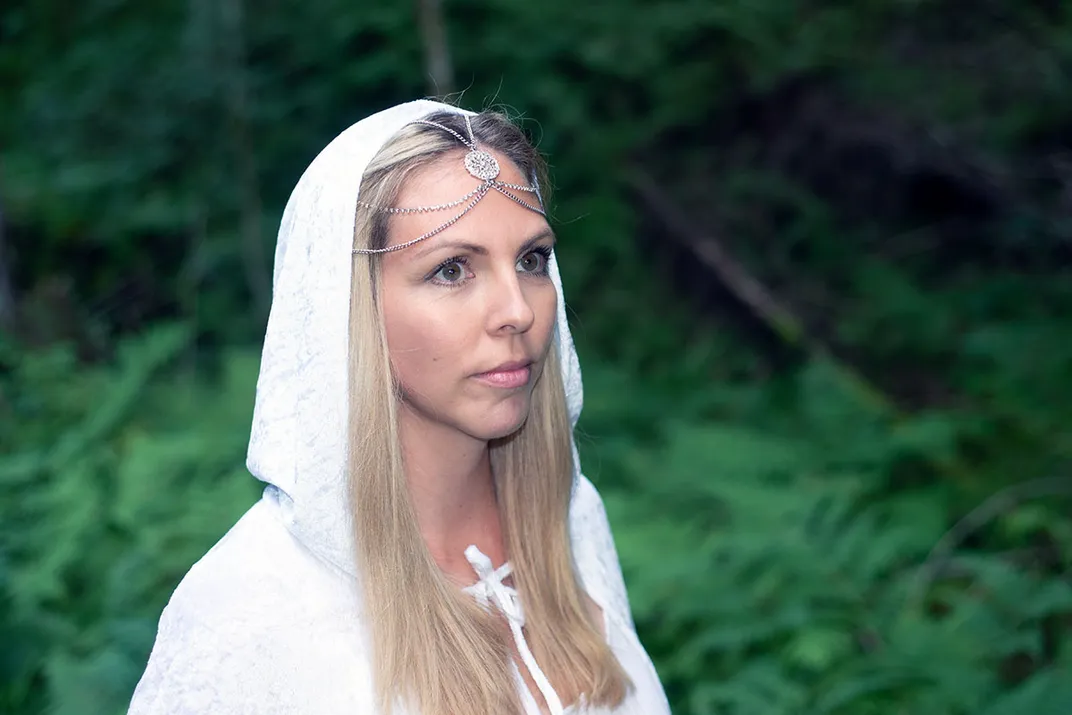 A person wears a white robe and head covering with silver veil in the woods.
