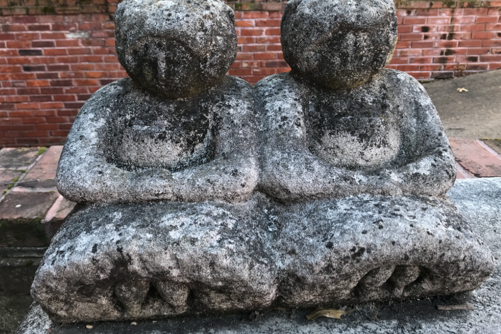 A sculpture of two seated women, covered in moss, dirt and weathered with time, rests outside in front of a sidewalk and brick wall