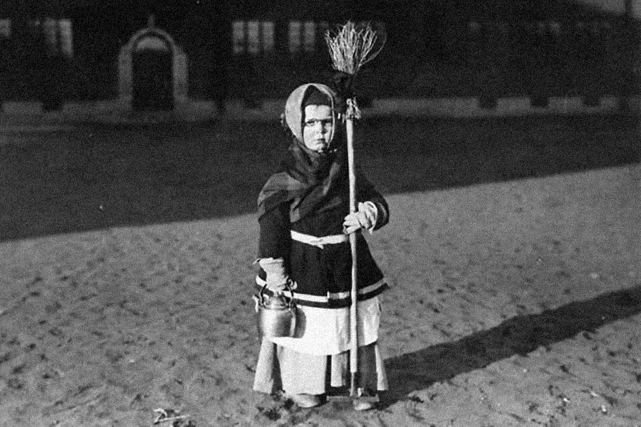 A young girl poses outside a building, wearing a dark coat, layers of skirts, and a kerchief over her hair, holding a broom in one hand and a kettle in the other. Black-and-white photo.