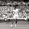 Althea Gibson in mid-swing on a tennis court, positioned in an athletic stance with her racket extended. A large crowd of spectators fills the stadium seating in the background.