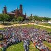 Thousands of people assemble on the green grass of the National Mall to form a sunburst shape, the Smithsonian logo. The red-brick Smithsonian Castle rises in the background.