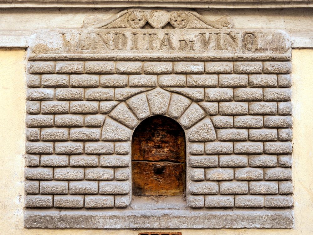 A small door cut into the side of a house. The door has engraved stone around its entrance, made to look like a tiny building facade. It bears an inscription that has worn away, but includes the word "VINO"