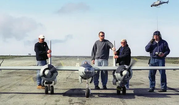Four men stand on a paved runway behind a large, grey twin-engine remote-controlled model airplane. The man on the far right holds a radio transmitter with a long antenna, while a small helicopter is visible in the sky behind him. The scene is set under