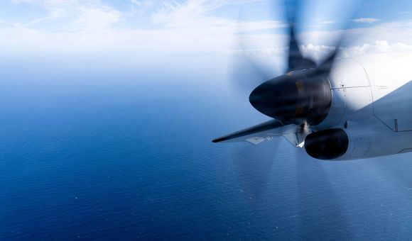 Wing view from a propeller plane flying over a vast ocean with a vibrant blue sky and clouds.
