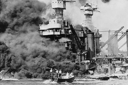 A small boat rescues a seaman from the 31,800 ton USS West Virginia burning in the foreground. Smoke rolling out amidships shows where the most extensive damage occurred.