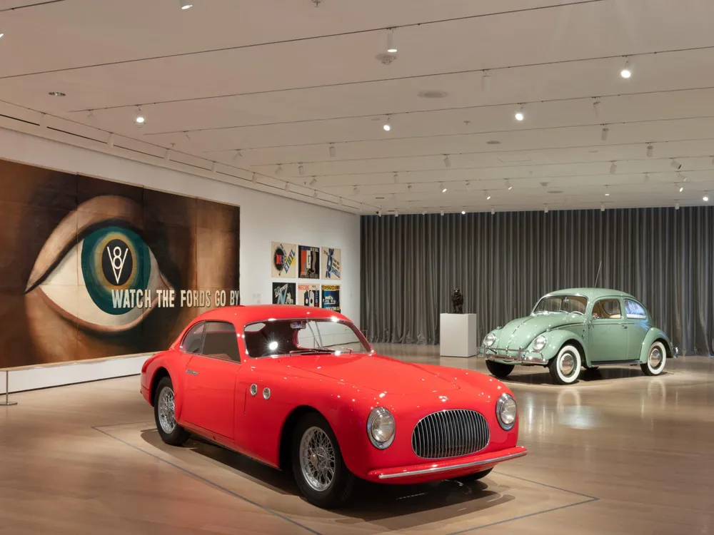 A view of a museum gallery with a bright red car on display in front of a light green Beetle; on the wall, an enormous lithograph of a human eye with the words Watch the Fords Go By