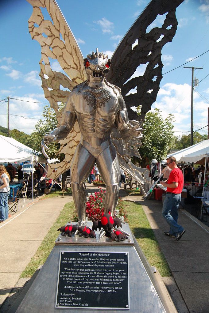 A statue of a silver humanoid moth with butterfly-like wings and red eyes, shown during daytime.