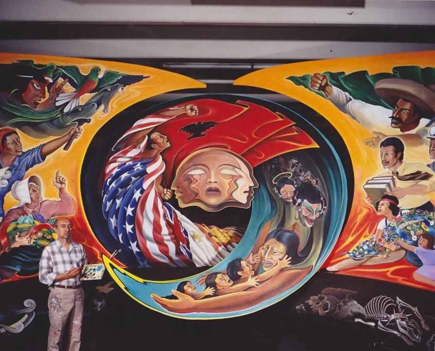 Leo Tanguma stands in front of a large mural-in-progress that features mythical figures, revolutionaries, laborers, and other figures of Mexican and Chicano history.