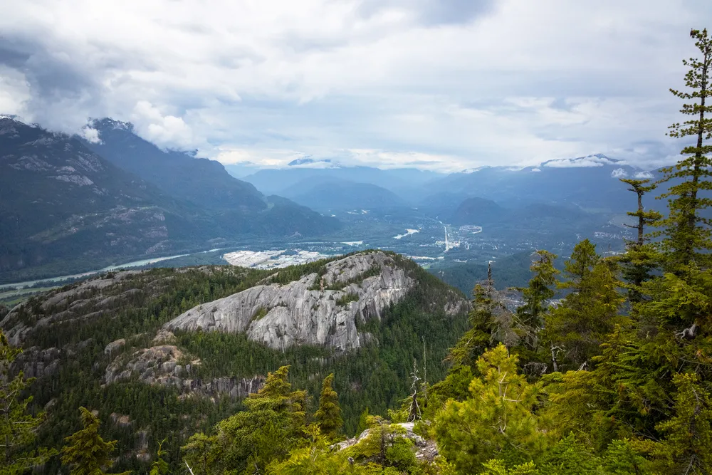 A view of the Stawamus Chief and the Squamish Valley, captured from the Sea to Sky Gondola's Panorama Trail. The wide angle perspective captures the granite monolith and the vast, forested landscape leading down to the town in the distance.