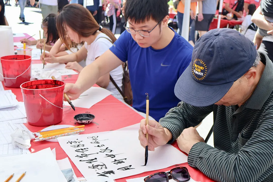 People of all ages sit together at an outdoor table painting calligraphy with brushes and buckets of black ink.