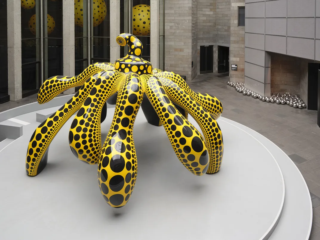 Yellow and black sculpture with legs on a white pedestal inside a museum