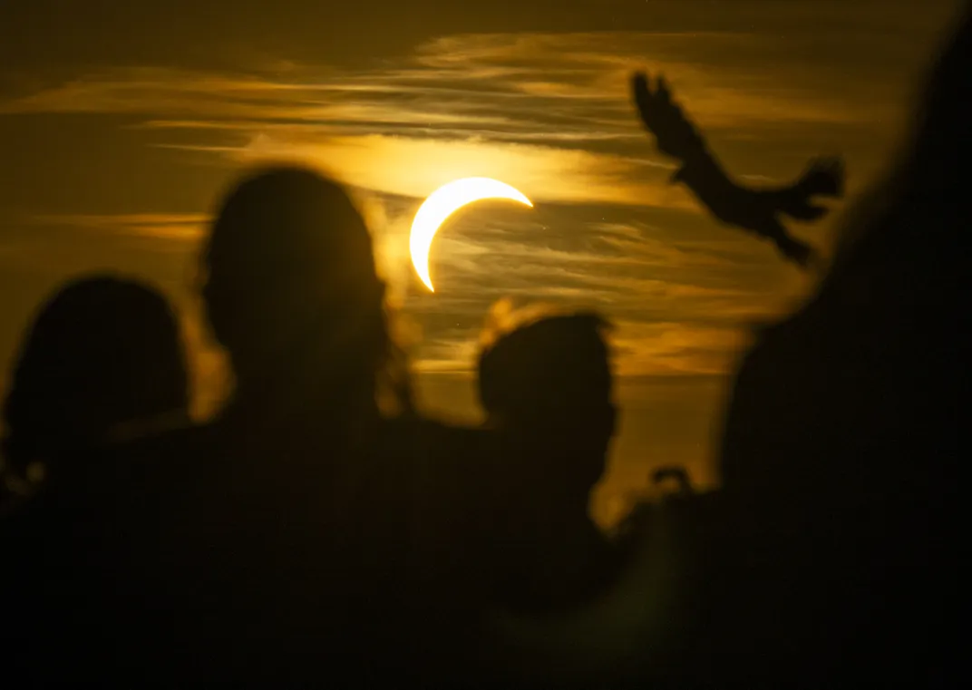 a partially eclipsed sun is behind clouds as silhouettes of humans and a flying bird are in the foreground
