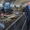 A white man in his mid-50s stands on a mezzanine overlooking several military jet airplanes parked on the concrete floor of a large hangar. The man is smiling and wearing a dark gray suit, blue shirt, and tie.