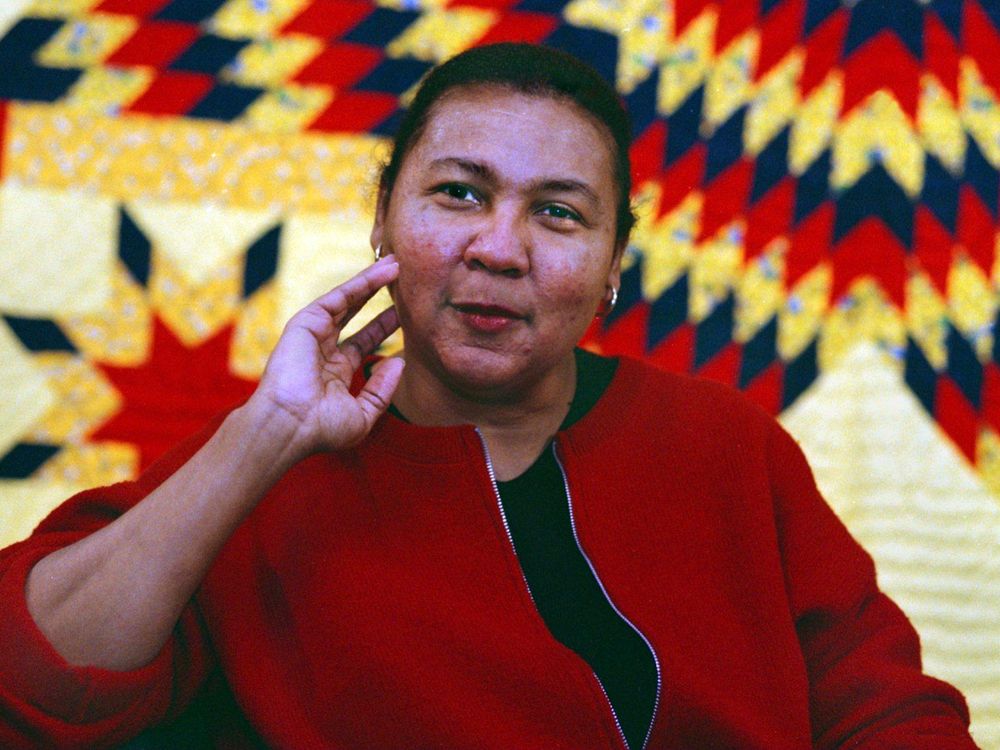 hooks, a Black woman in a red blazer, sits in front of a red, yellow and black quilt with her hand resting lightly on her chin and a small smile