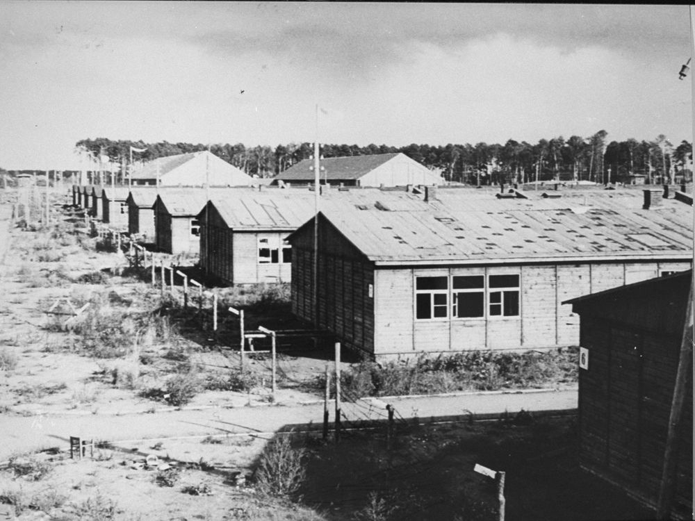 A black and white image of low rectangular buildings with peaked roofs, grassy dirt ground, black windows and shadows