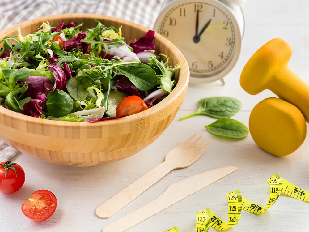 Wooden bowl of salad on a table with a clock, some utensils, a curled tape measure and barbells