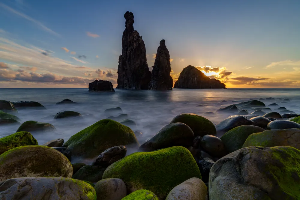 Sunrise light filters behind the sea stacks of Ribeira da Janela in Madeira, illuminating the moss-covered rocks as waves gently wash over them. A moment that blends rugged coastline, soft motion, and warm morning glow.