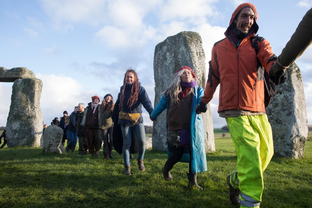A group of people hold hands and smile, appear to be moving in a circle through and around the Neolithic stones