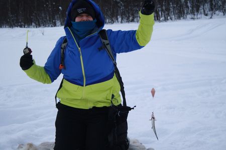 Person wearing winter gear kneeling in the snow holding up a fishing line with a small fish hooked on it