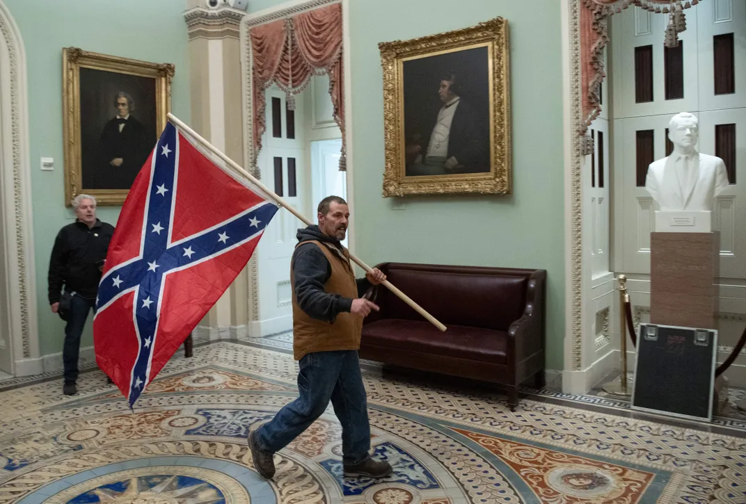 A white man wearing a hoodie walks upright, carrying a huge Confederate flag on a pole over his shoulder, through the halls of the Capitol building. Behind him, two gilded portraits of white senators, and a white man dressed in black standing behind him.