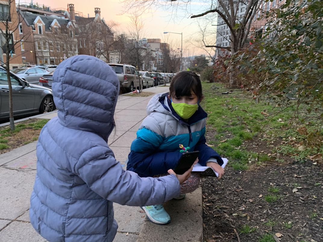 Two kids using a notebook and cell phone while outside.