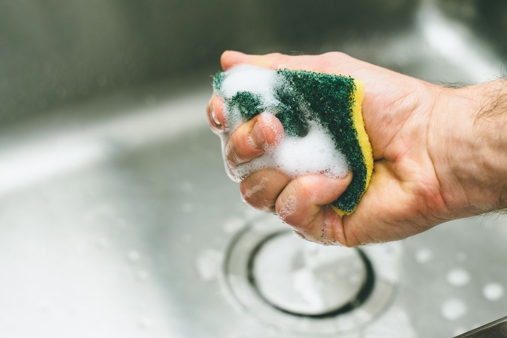 Image of a hand wringing out a soapy green and yellow kitchen sponge into a sink