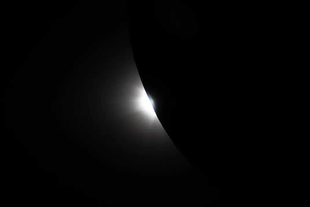 the bright orb of the sun emerges from behind the darkened moon, looking small compared to the moon's immensity