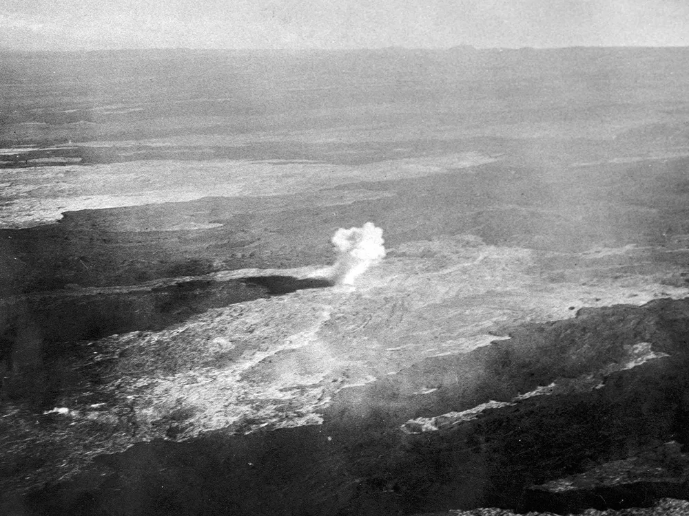 An old photo, tinted brown, depicts a flat barren landscape with a single pillar of dense white smoke at the center of the photo.