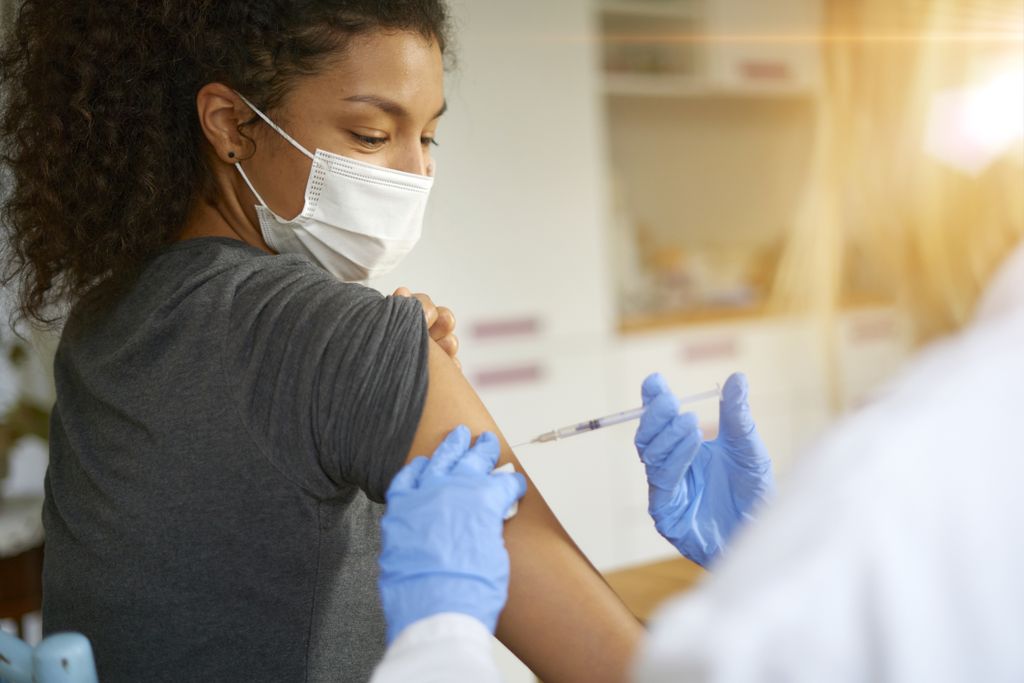 a woman in a mask gets a vaccine in her arm from a person in a lab coat and gloves