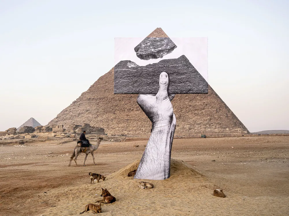 A disembodied black and white hand emerges from the sand and holds a photograph, which creates the effect that the tip of a pyramid behind is separated and floating above its base