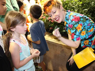 A woman wearing a brightly colored shirt with flowers smiles while showing two children a butterfly that is perched on her finger.