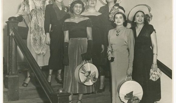 A black-and-white photograph of seven women standing on a flight of stairs. Mae Reeves stands to the farthest right on the lowest step and wears a white hat and floral patterned handbag.