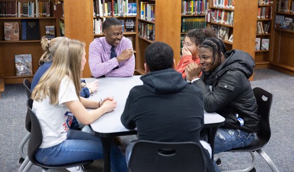 Teacher Kurt Russell sits at a square table in the school's library with five students, smiling and in conversation.