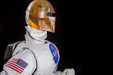 Photographed against a black background is a robot in profile wearing a silver uniform and golden-colored helmet.