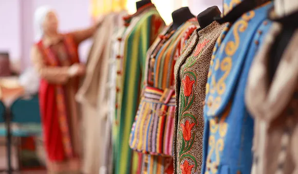 In the foreground, a lineup of mannequins each with colorful embroidered garments. At the end of the line, out of focus, a woman dresses a mannequin.