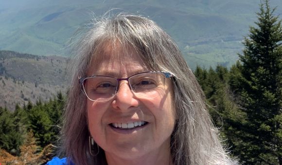 Head and shoulders photo of Denise Breitburg, a woman with glasses and shoulder-length gray hair, with forested mountains in the background.