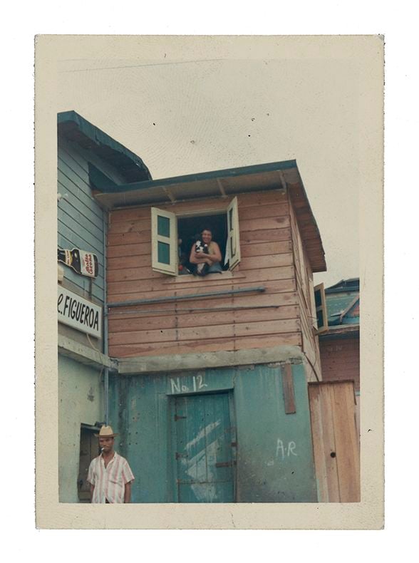 Photograph of a wooden house painted blue with red shutters. A woman leans out of an upstairs window holding a dog and a man is below on the street smoking a cigarette.