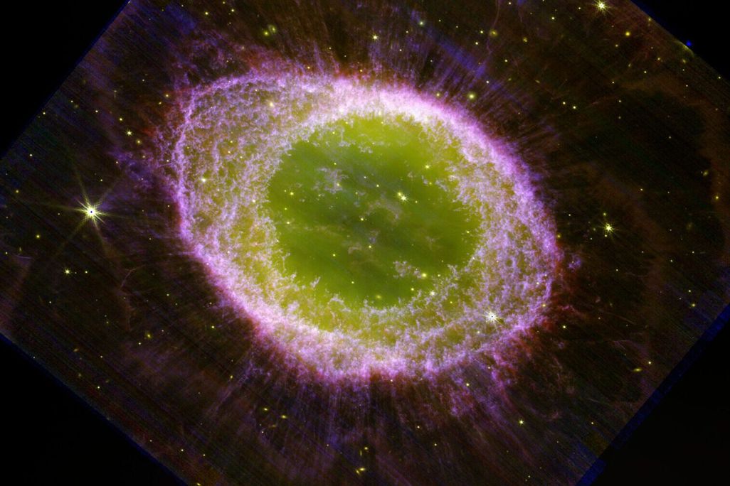 An image of the ring nebula, with a large green center, a purple outer ring, and wisps of purple beyond that. Stars and other galaxies glow yellow in the background.