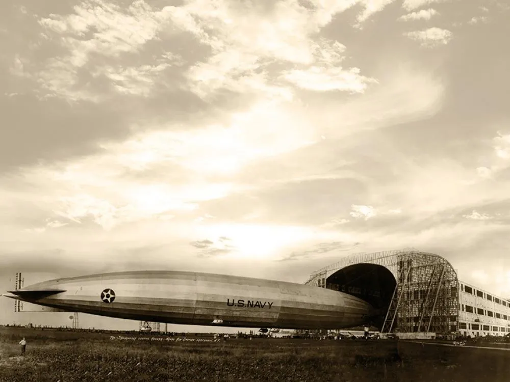 A black-and-white photo, tinged light brown, shows around 3/4 of massive airship as it exits a pitch black hangar opening.