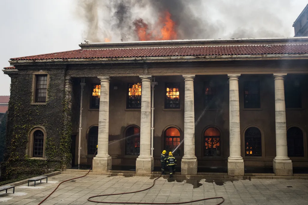 Two firefighters in gear, looking small compared to tall white columns, aim jets of water at the windows of a historic building; orange flames leap from the windows and ceiling of the structure and smoke pours out its top