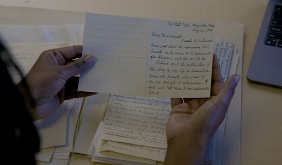 Two hands holding a card with handwritten information. A stack of other handwritten papers is visible on the table below.