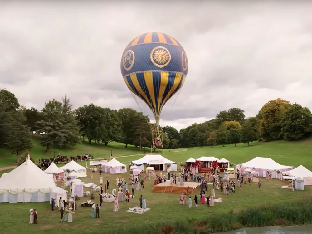 A vibrant hydrogen balloon with intricate blue and gold designs floats above a bustling outdoor event set against a cloudy sky. Below, several white tents are set up in a grassy park, with groups of people mingling and walking around.