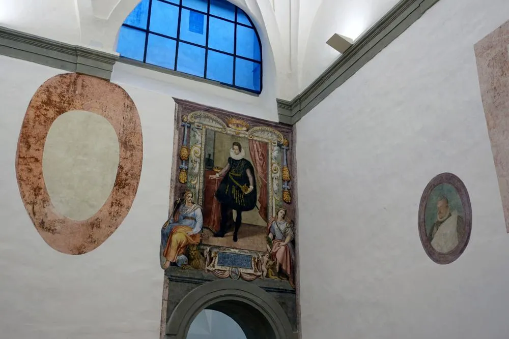 A white plaster-walled room with one archway, over which a large square fresco hangs, depicting a man in black garb standing in front of a luxurious background