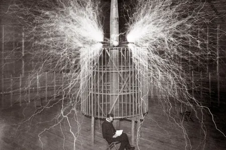 Both genius and impresario, Serbian-American inventor Nikola Tesla reads in his remote Colorado Springs laboratory in 1899 next to a magnifying transmitter that generates millions of volts of electricity. While far too dangerous to sit near—the image is a double exposure—his gigantic Tesla coil created the first human-made lightning.
