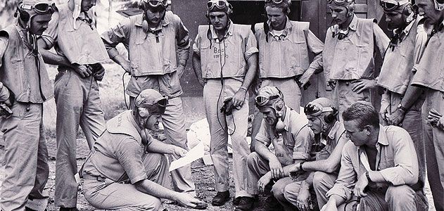 At the Black Sheep Squadron's base on the South Pacific island of Espiritu Santo, Boyington (holding paper) briefs his pilots on an upcoming mission.