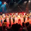 Performers wave, forming a ring around a red, circular stage, with a packed audience and rigging hanging above.