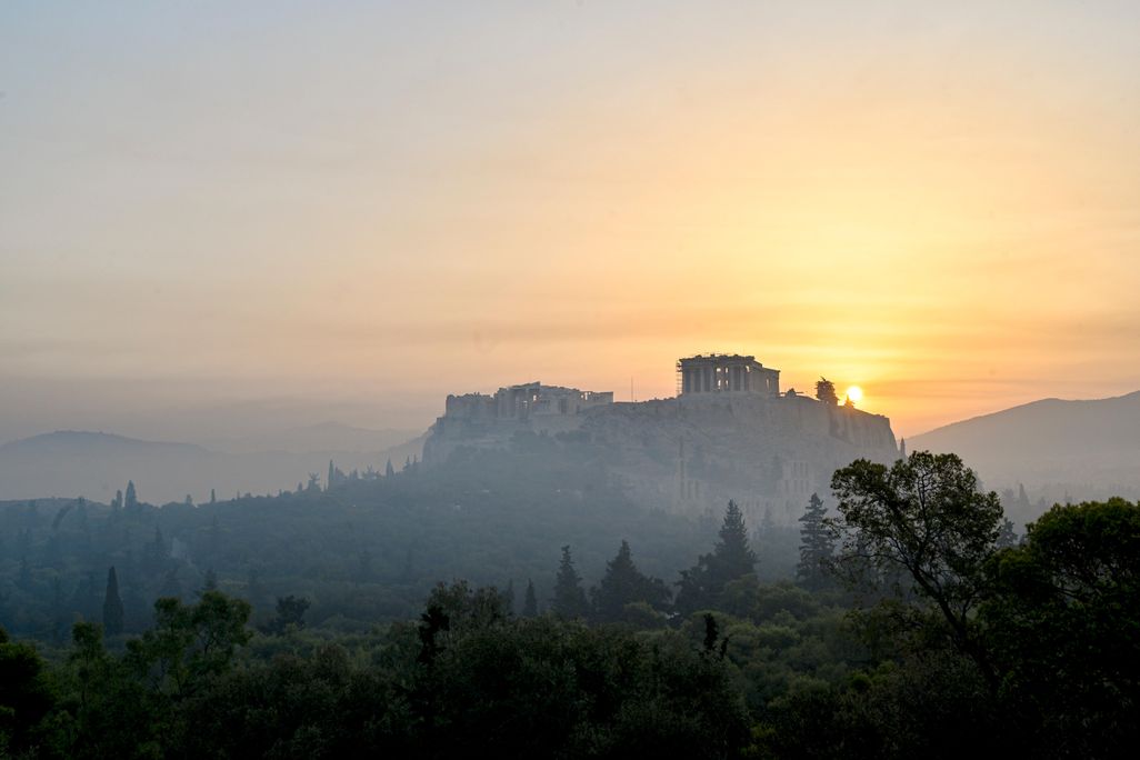 A view of the Parthenon in the distance on a hilltop, with the sun low in the sky behind a haze of smoke